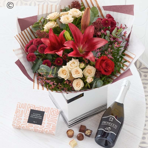 blooms in red and green, with peach and cream highlights and pops of burgundy and cerise, a bottle of Prosecco and a box of chocolates