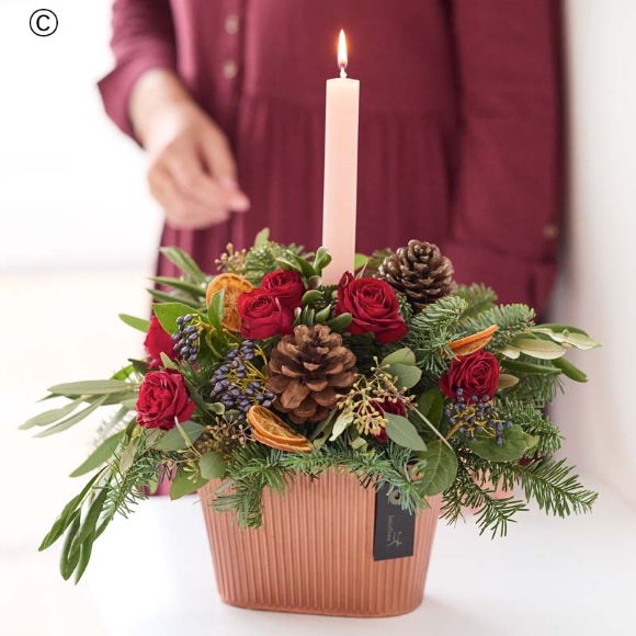 arrangement centred around a festive candle with blooms in red and green, and peach and cream highlights and pops of burgundy and cerise