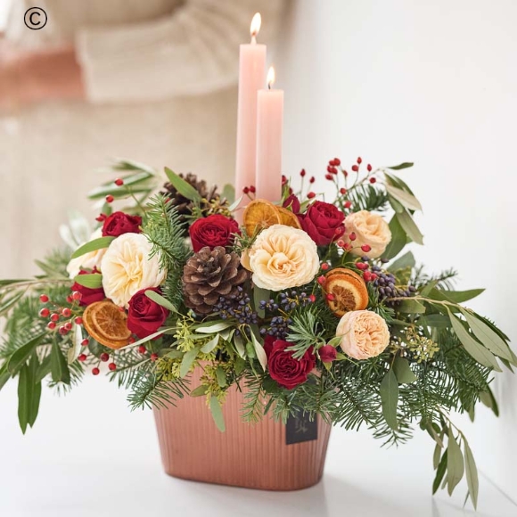 an arrangement centred around two festive candles featuring blooms in red and green, with peach and cream highlights and pops of burgundy and cerise