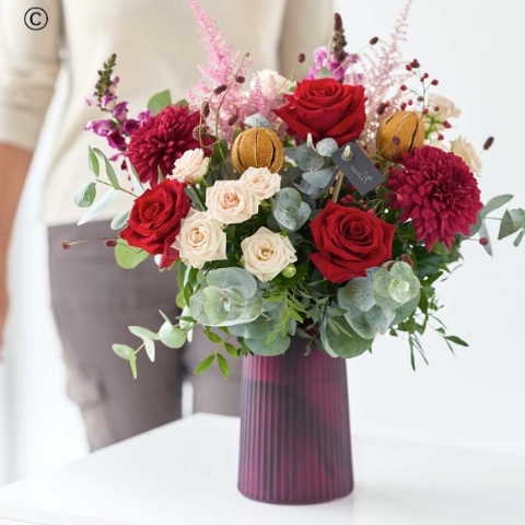 transparent ribbed handblown vase filled with festive blooms in red and green, with peach and cream highlights and pops of burgundy and cerise